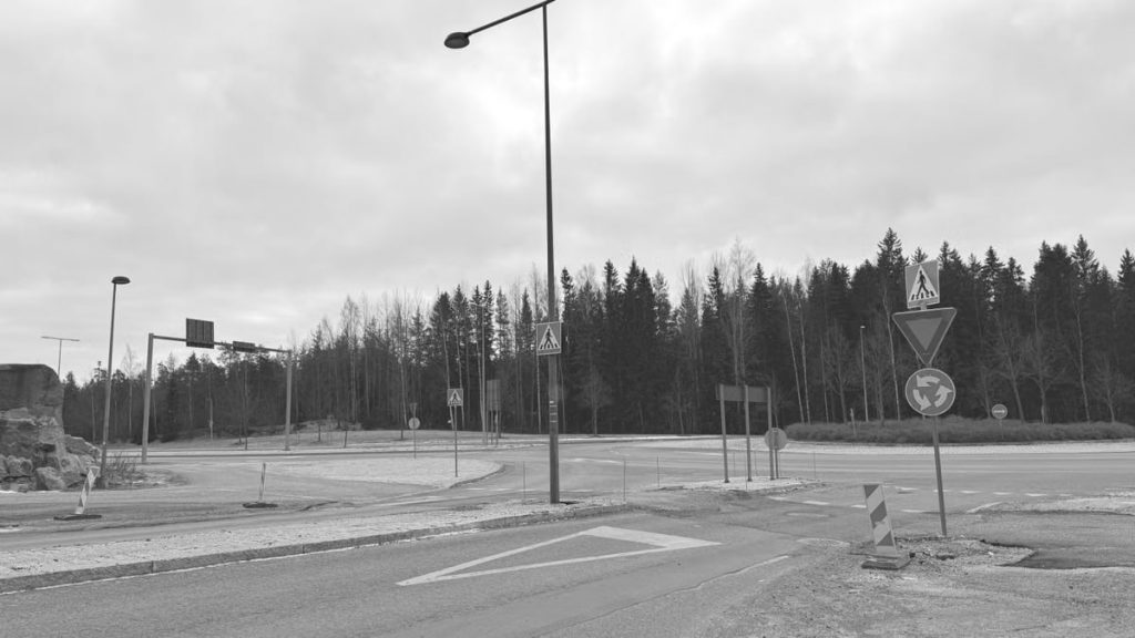 Black and white image of an empty roundabout with traffic signs and trees in the background.