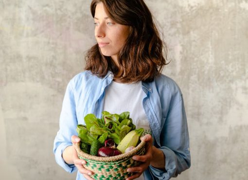 A Caucasian woman gazes away while holding a basket filled with fresh vegetables. Indoor setting.